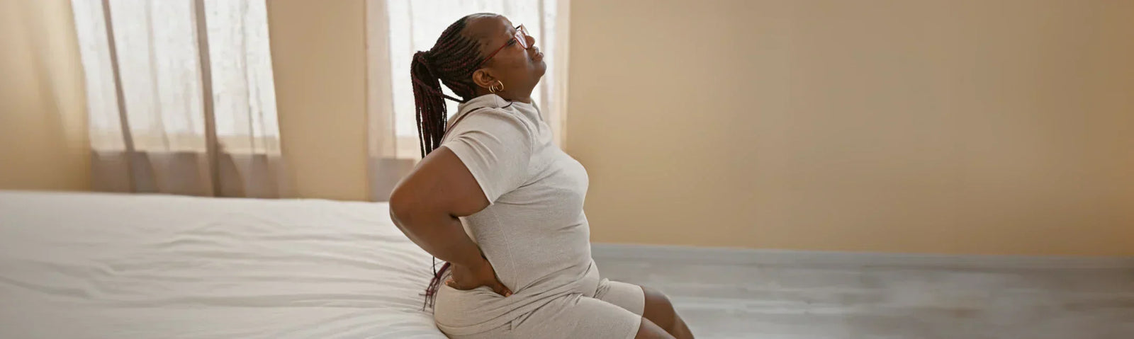 woman sitting on the edge of a mattress, with her arms bent and holding her lower back in pain