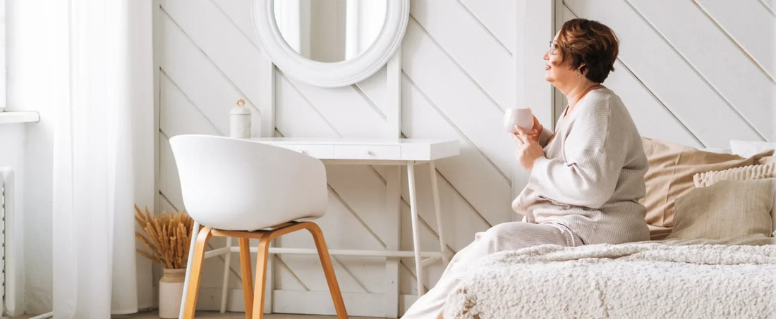 woman sitting on edge of a mattress, holding a cup of coffee staring out a window
